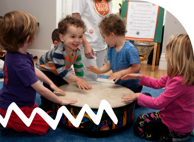 Smiling toddlers gathered around and playing a large drum. An adult wears a white and red "Little Roots" t-shirt.