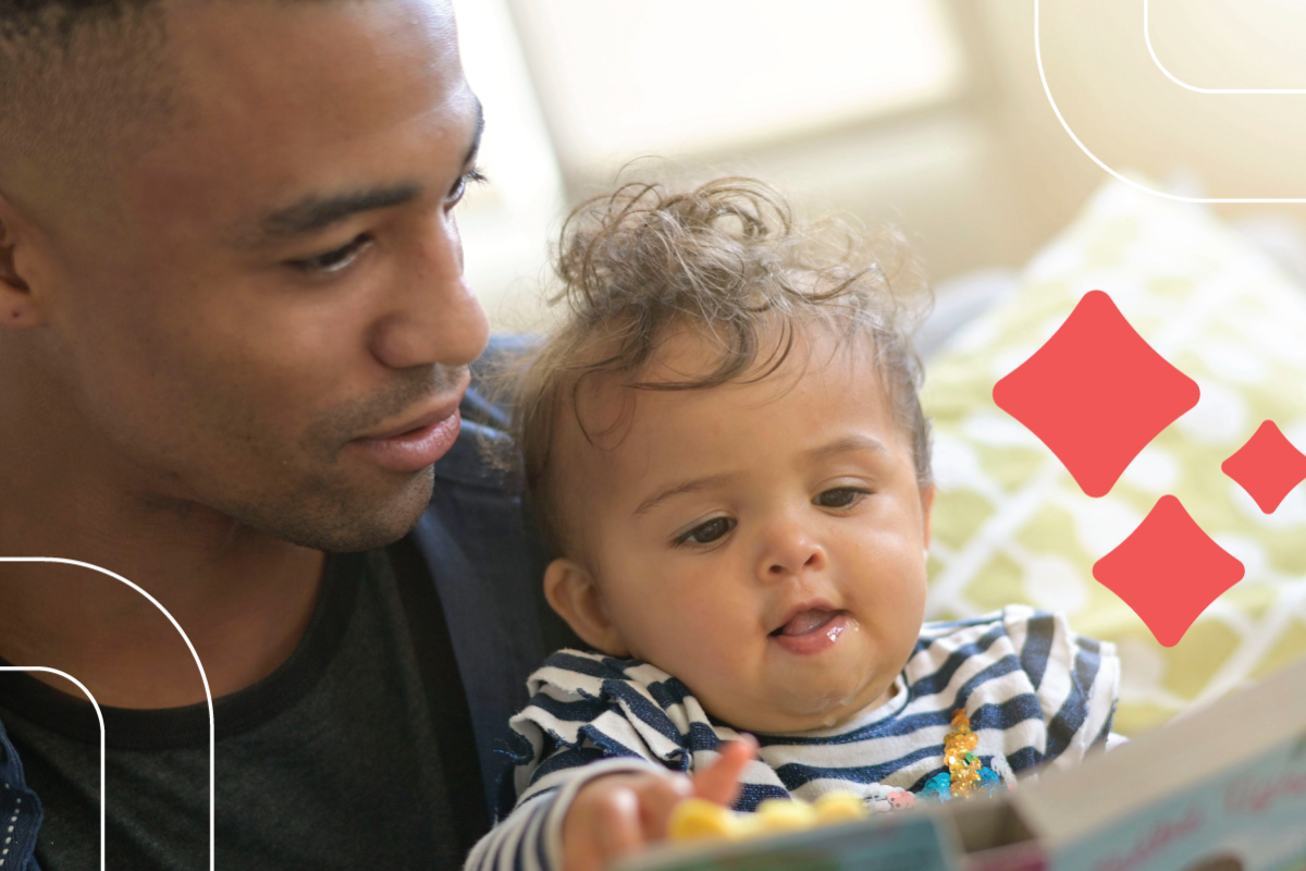 a baby reaches for a picture book from the lap of their dad