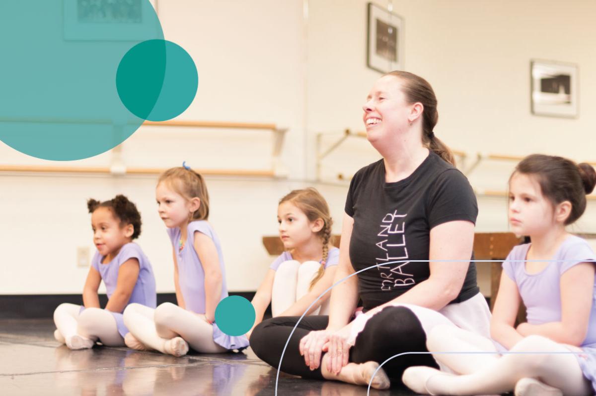 a ballet instructor sits on the floor with four young dancers in tutus 